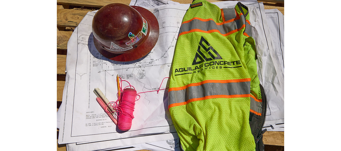 A red hard hat, neon safety vest with the Aguilar Concrete Services logo, pink string, and markers rest on top of construction blueprints laid out on a wooden surface at a job site.
